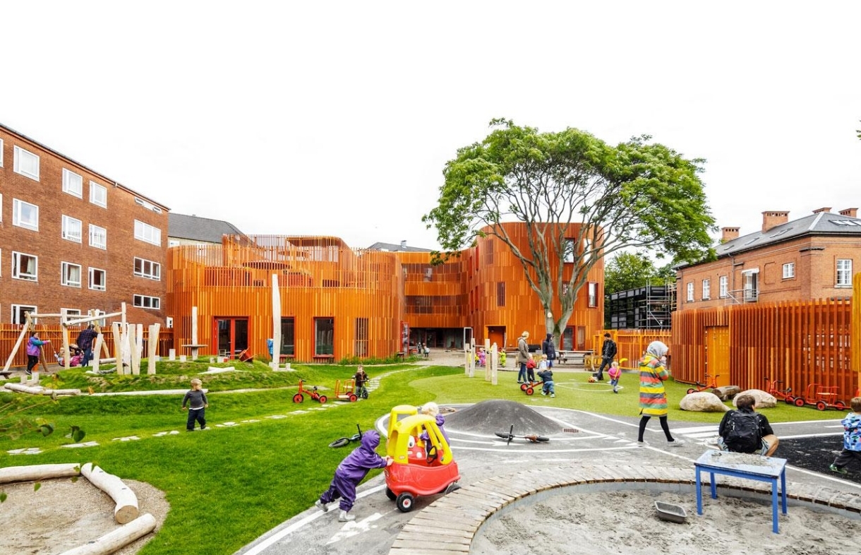 Pátio de creche dinamarquesa com arquitetura em madeira laranja e crianças brincando ao ar livre